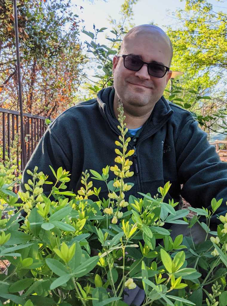 Photo of Indigo Underwood outside in a garden, sitting behind some bright green plants.