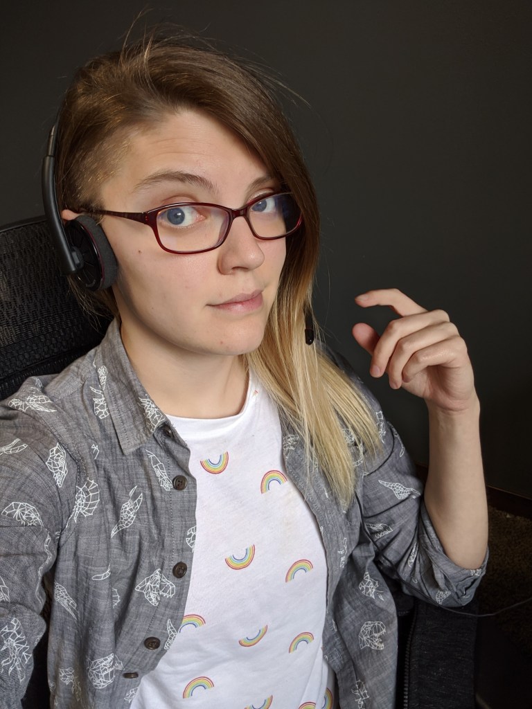 Photo of Galacia wearing a white shirt with rainbows on it and a button-up gray shirt over it. Galacia has headphones on and glasses.