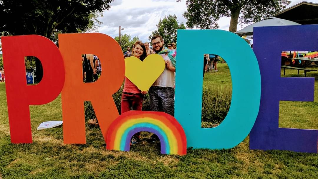 Photo of two people stand behind a wooden painted rainbow and hold a large yellow-painted heart. They form the I in the word "pride"as they stand in-between giant adult person-sized letters P and R on one side and D and E on the other side. These giant letters are set up in a grassy park with many people in the background.