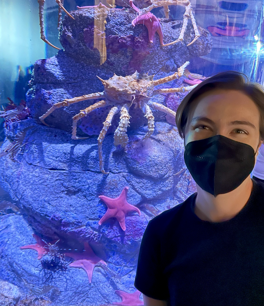 Image of the author, Dr. Antell, at the Aquarium of the Pacific, California. The background is a purple rock behind aquarium glass, on which three purple starfish and some large crabs climb. Dr. Antell has short, brown hair and is wearing a black mask - but you can tell that Dr. Antell is smiling by the eyes. Dr. Antell wears a black t-shirt.