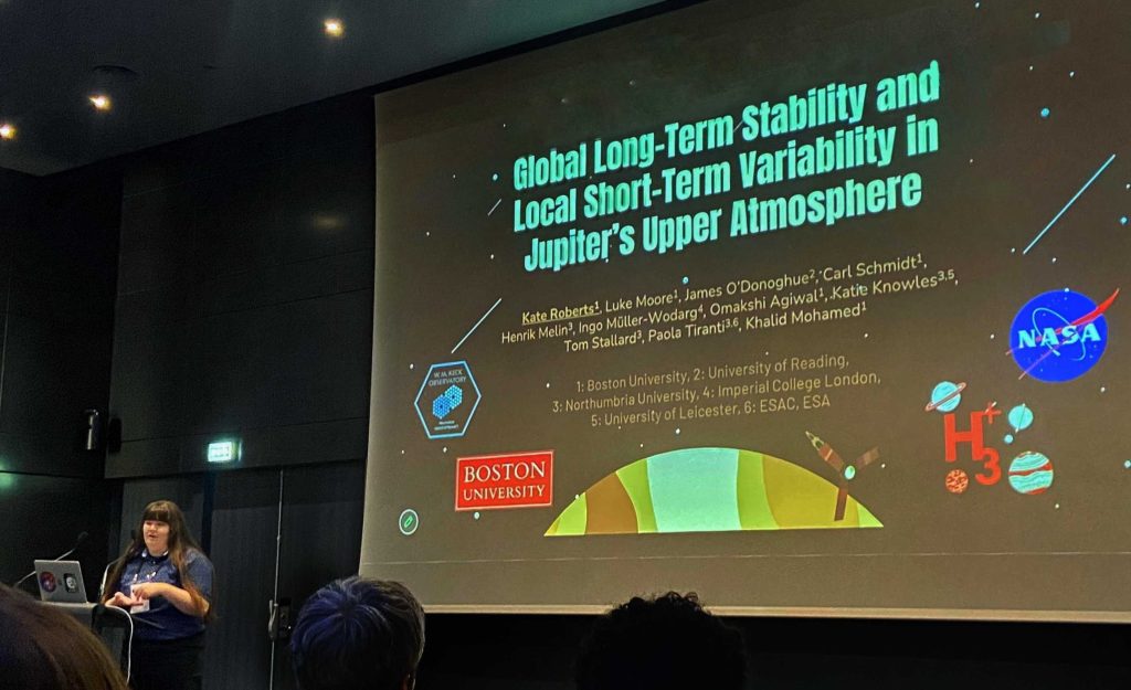 A person with a laptop stands in front of a presentation titles Global Long-Term Stability and Local Short-Term Variability in Jupiter's Upper Atmosphere. 
